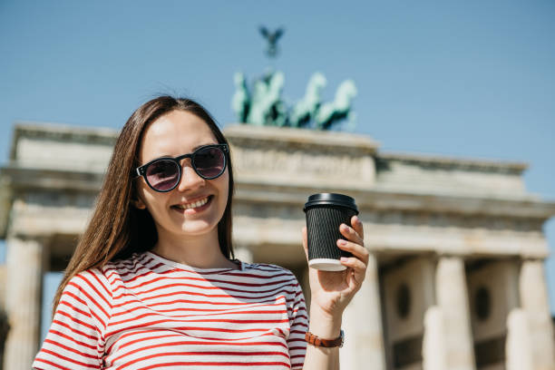 A person holds a disposable cup with coffee or another drink on the background of the Brandenburg Gate in Berlin.