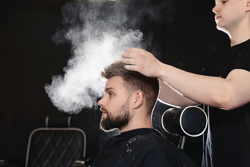Professional hairdresser barber using talcum powder for his client hair sitting in a barber chair on black background