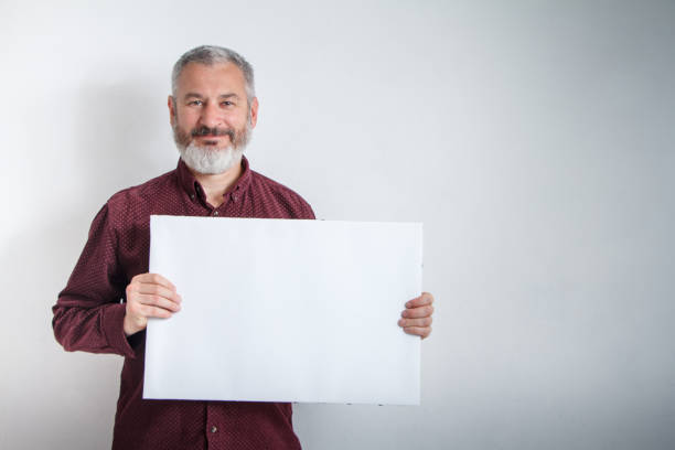 istock Gray-haired man with a beard holding white empty signboard with space for text in front of him 1150114272