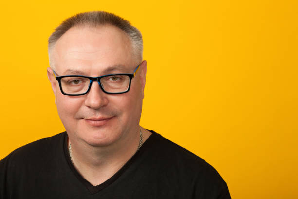 portrait de studio de l’homme de calvitie de 50 ans dans des verres, et t-shirt noir sur le fond jaune - 45 49 years photos et images de collection