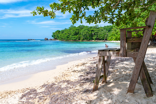 Wooden lifeguard chair on beautiful white sand turquoise blue beach on tropical Caribbean island. People enjoying ocean/sea water with lush trees and large rock formations in background on summer day.