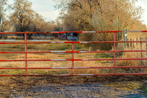 No Trespassing sign on the gate of a private property viewed on a sunny day. A grassy terrain with trees against cloudy sky can be seen behind the weathered red gate.