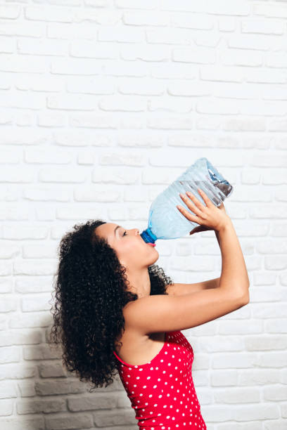 健康のためにペットボトルから水を飲む喉の渇いた若い女性 - ガロン ストックフォトと画像