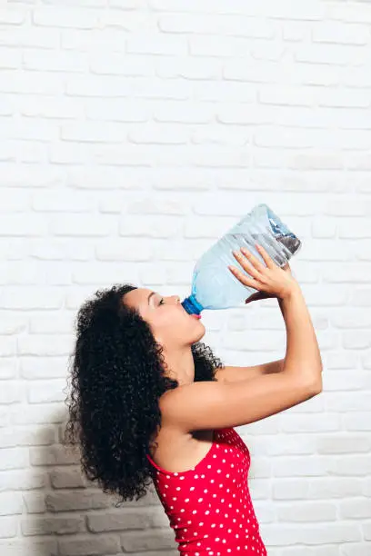Portrait of happy african american woman drinking mineral water from plastic bottle. Thirsty black girl with drink container in summer.