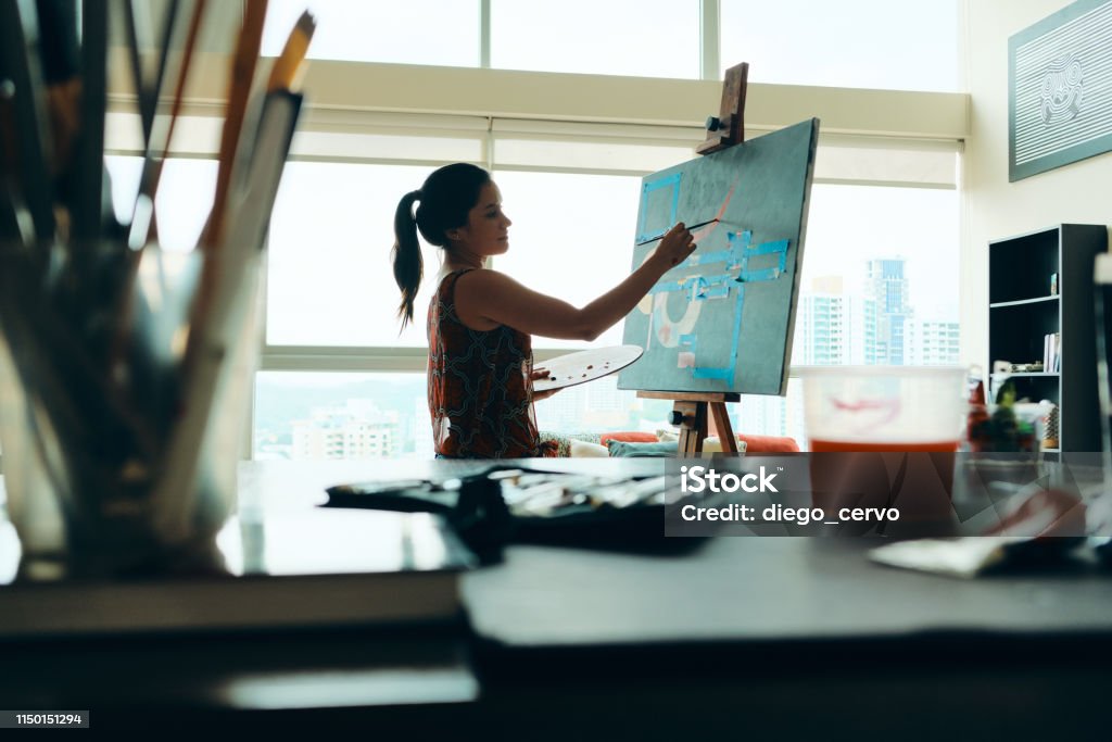 People Girl Woman Painting In Lab For Arts Hobby Work Portrait of young latino woman painting for hobby in her home studio, practicing with paint, brushes, easel and colors. Painting - Art Product Stock Photo