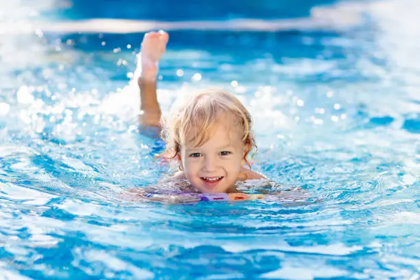 Child learning to swim in outdoor pool of tropical resort. Kids learn swimming. Exercise and training for young children. Little boy with colorful float board in sport club. Swimming baby or toddler.