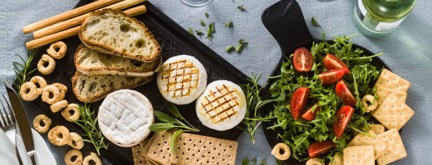 banner of grilled Italian Tomino cheese served on a table with white wine, crackers, grissini and taralli with aromatic herbs and arugula and tomato salad on a blue linen festive tablecloth. summer menu