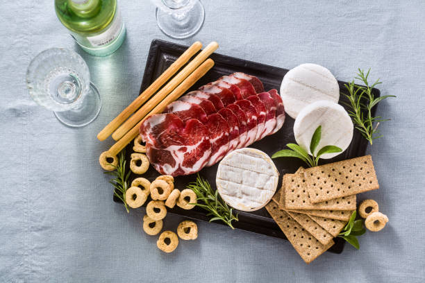 冷たいカットとチーズは、白いワイン、クラッカー、グリッシーニ、タラーリのテーブルの上に、青いリネンのお祝いのテーブルクロスの上に芳香族のハーブと一緒に出されます。 - tomino ストックフォトと画像