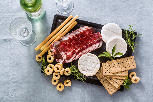 cold cuts and cheese are served on a tray on a table with white wine, crackers, grissini and taralli with aromatic herbs on a blue linen festive tablecloth.