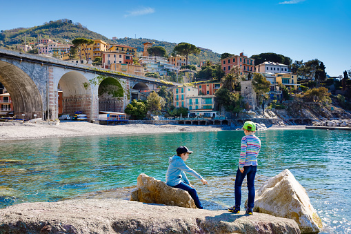 Two little kids boys climbing on beach stones of Mediterranean sea in Liguria region, Italy. Siblings brothers in Zoagli, Cinque Terre. Beautiful Italian city with colorful houses. Family vacations
