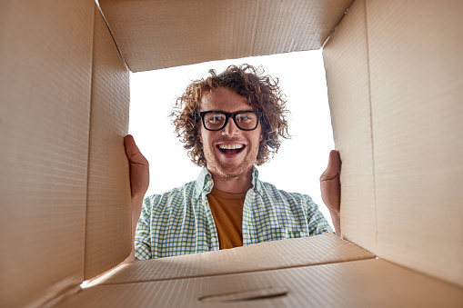 From below attractive young man in stylish glasses looking inside open box with belongings while relocating in new place