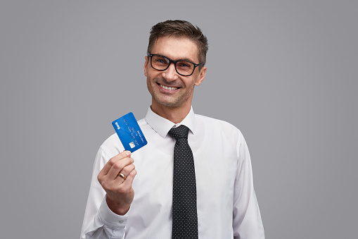 Excited male in elegant shirt and stylish glasses cheerfully smiling and showing credit card while standing on gray background and looking at camera