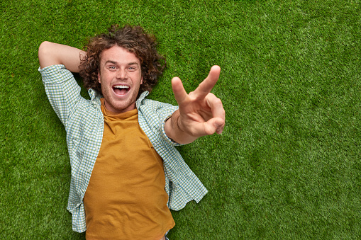From above excited guy with curly hair showing V sign and looking at camera while lying on green lawn