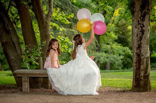 Two girls dressed in communion playing in a park in Madrid with colorful helium balloons surrounded by trees and nature