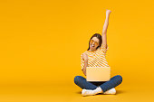 Winner! Excited smiling girl sitting on floor with laptop, raising one hand in the air is she wins, isolated on yellow background