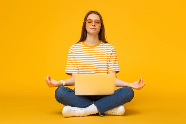 junge frau, die auf dem boden mit laptop meditiert in yoga-lotus posieren isoliert auf gelben hintergrund - floor women sitting yoga stock-fotos und bilder