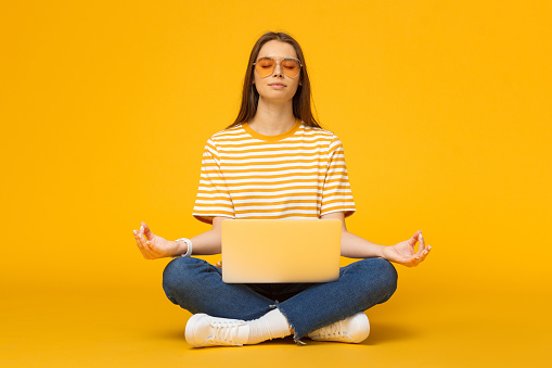 Mujer joven sentada en el piso con Laptop meditando en yoga de loto pose aislada sobre fondo amarillo photo