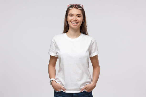 junge lächelnde frau steht mit händen in taschen, trägt blankes weißes t-shirt mit kopierplatz für ihr logo oder text, isoliert auf grauem hintergrund - artificial model fotos stock-fotos und bilder