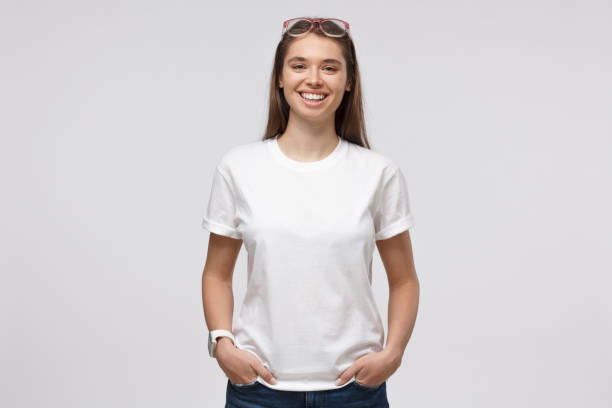 Young smiling woman standing with hands in pockets, wearing blank white t-shirt with copy space for your logo or text, isolated on gray background Young smiling woman standing with hands in pockets, wearing blank white t-shirt with copy space for your logo or text, isolated on gray background caucasian appearance stock pictures, royalty-free photos & images