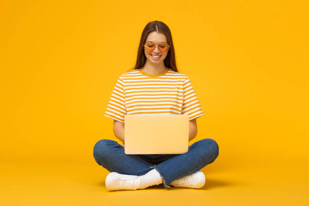 istock Smiling girl sitting on floor, using laptop, isolated on yellow background 1150254178
