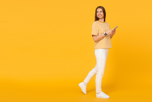 Young caucasian woman walking and communicating via phone, isolated on yellow background with copy space on left