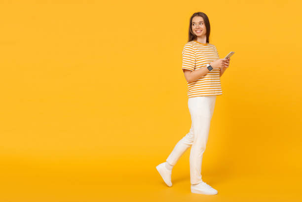 young caucasian woman walking and communicating via phone, isolated on yellow background with copy space on left - full length imagens e fotografias de stock