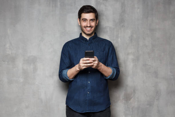 istock Young man looking at camera with smartphone in hands, isolated on gray textured wall background 1150254229