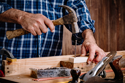 Close-up of Mature carpenter using hammer and nail on wood plank at home workshop