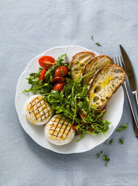 grilled Italian Tomino cheese served on a table with arugula salad and fresh homemade ciabatta bread and tomato on a blue linen festive tablecloth. summer menu