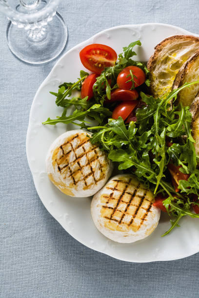 grilled Italian Tomino cheese served on a table with arugula salad and fresh homemade ciabatta bread and tomato on a blue linen festive tablecloth. summer menu
