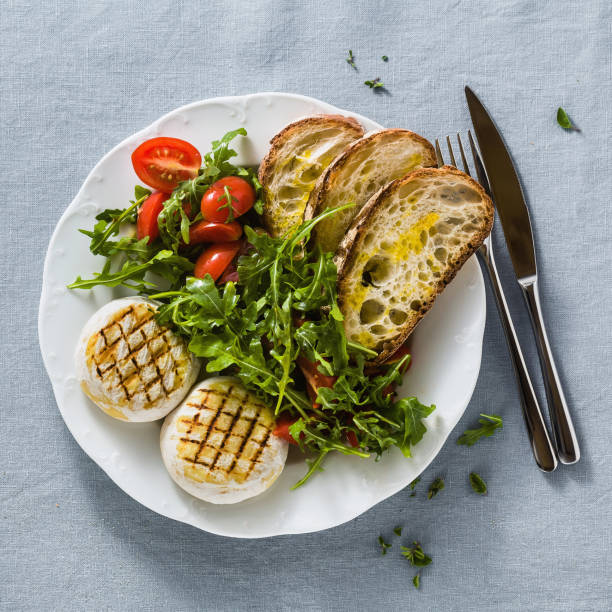 grilled Italian Tomino cheese served on a table with arugula salad and fresh homemade ciabatta bread and tomato on a blue linen festive tablecloth. summer menu