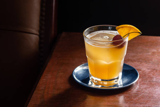 Amaretto Sour Cocktail on the Rocks with Cherry and Orange Garnish in Dark Luxurious Bar An Amaretto Sour cocktail on the rocks with a cherry and orange slice garnish on a dark wood table next to a leather seat. This drink is made with amaretto liqueur, lemon, and simple syrup (or sour mix) and topped with a cherry and orange. Taken in a dark luxurious bar or restaurant. sour taste stock pictures, royalty-free photos & images