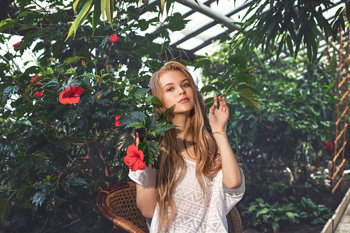 Young beautiful happy blonde girl model with red flowers on tropical plants background in greenhouse