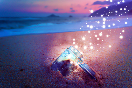 Colorful magic glowing light bugs leaving mason jar washed ashore of beautiful sunset beach at evening.