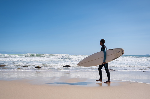 Mid distant view of an African teenage boy carrying surfboard during weekend. Full length of surfer is walking on sand. He is spending time doing activities at beach.