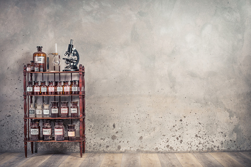 Old laboratory bottles, antique microscope, vintage lab glassware support on the wooden shelving front concrete wall background. Retro style filtered photo