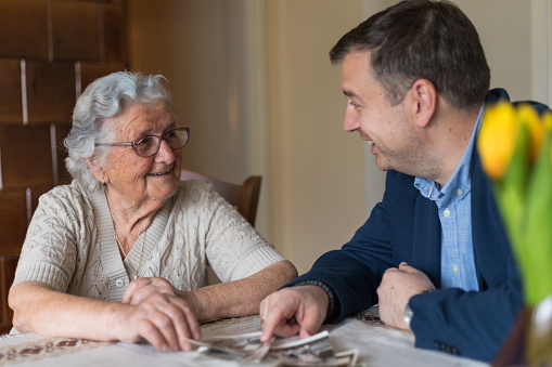 Senior woman with her adult grandson are looking old photos and remember the old memories.Man enjoys the company of his grandmother and spending wonderful time with her.\nOld woman with gray hair, glasses and a middle-aged man in a jacket are sitting at the table and looking at the photos in the old family house.