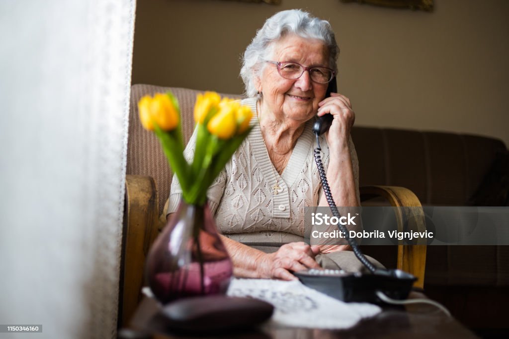 Happy senior woman talking on the phone in living room. Old woman with gray white hair and glasses sitting in her armchair in her home and talking on the phone. Grandmother is happy to talk to her children and grandchildren. Senior Adult Stock Photo
