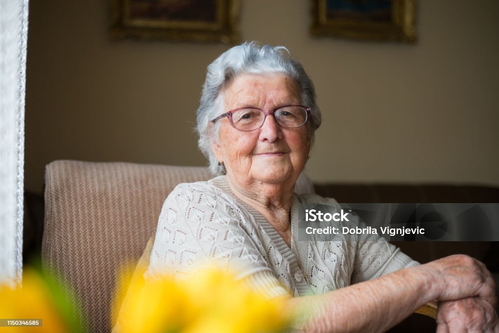 Close-up portrait of happy senior woman portrait Portrait of a beautiful old woman with gray hair and glasses is sitting in a chair in her home. Senior Adult Stock Photo