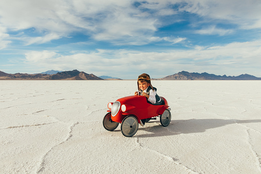 A young boy and businessman dressed in a business suit drives his vintage toy car on the salt flats located in Utah, USA. He is excited to race his business into profitability to the next travel destination.