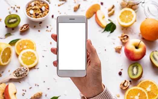 Female hands holding smartphone on healthy food background, woman using phone search mobile apps with diet nutrition plan cooking, vegan fruit granola seeds on white table, top view, mock up screen