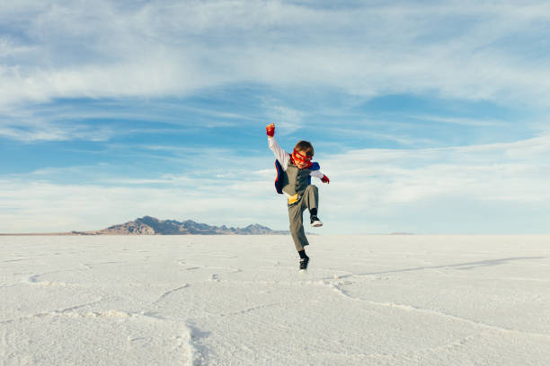 A young boy businessman is dressed up as a superhero with cape and jumps and flies through the sky while on the Bonneville Salt Flats in Utah, USA. This young entrepreneur is ready to take on challenges and lead his business into profitability.