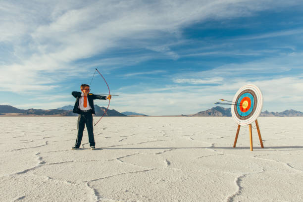 Successful Business Boy with Arrows in Target A boy businessman stands next to his arrows in a target, hitting their mark in the bulls-eye. Precision is paramount in target shooting and he has mastered it. The entrepreneur shows his preparation, determination and accuracy it takes to reach a goal. Image taken on the salt flats of Utah, USA. hit the nail on the head stock pictures, royalty-free photos & images