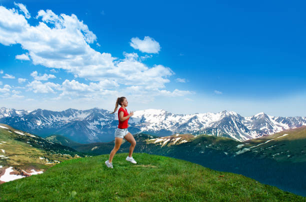 Young woman running along mountains. Smiling girl exercising on vacation trip. Snow capped mountains. View from Trail Ridge Road. Rocky Mountains National Park. Estes Park, Colorado, USA.