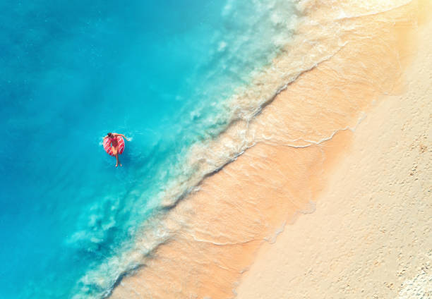 Aerial view of a young woman swimming with the donut swim ring in the clear blue sea with waves at sunset in summer. Tropical aerial landscape with girl, azure water, sandy beach. Top view. Travel Aerial view of a young woman swimming with the donut swim ring in the clear blue sea with waves at sunset in summer. Tropical aerial landscape with girl, azure water, sandy beach. Top view. Travel girls laying on beach stock pictures, royalty-free photos & images