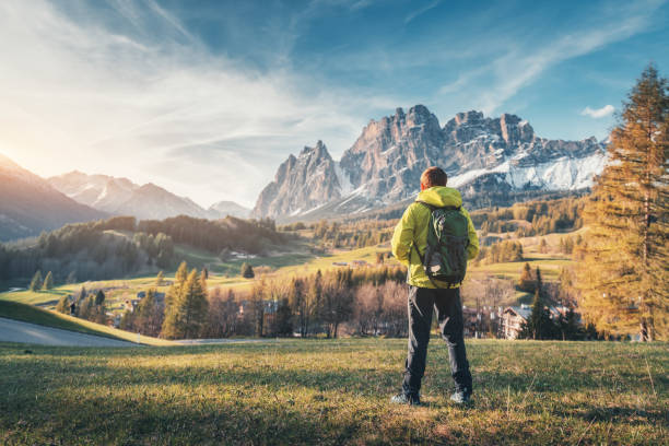 istock Young man in yellow jacket with backpack standing on the green meadow and looking on mountains at sunset in spring. Landscape with sporty guy, snowy rocks, trees, houses, blue sky in Italy. Travel 1150377021