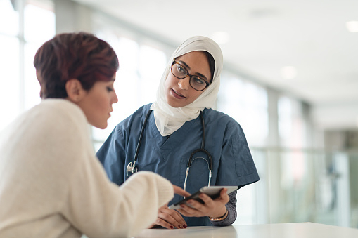A female doctor of Middle Eastern ethnicity is meeting with her female patient. The two women are seated at a table. The doctor is showing the patient test results on a digital tablet. The patient has her back to the camera.