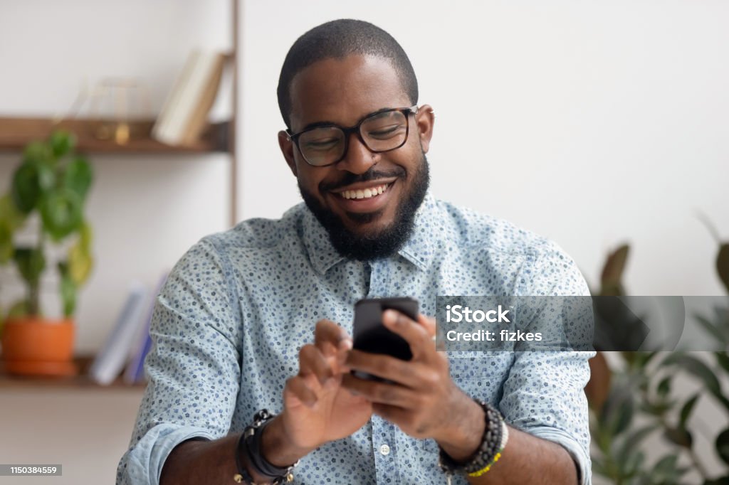 Happy african american businessman using phone mobile apps at workplace Happy african american businessman using phone mobile corporate apps at workplace texting sms, smiling black man looking at smartphone browsing internet, office technology and digital communication Customer Stock Photo