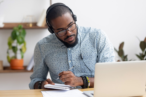 Enfocado hombre de negocios africano en auriculares escribiendo notas viendo webinar photo