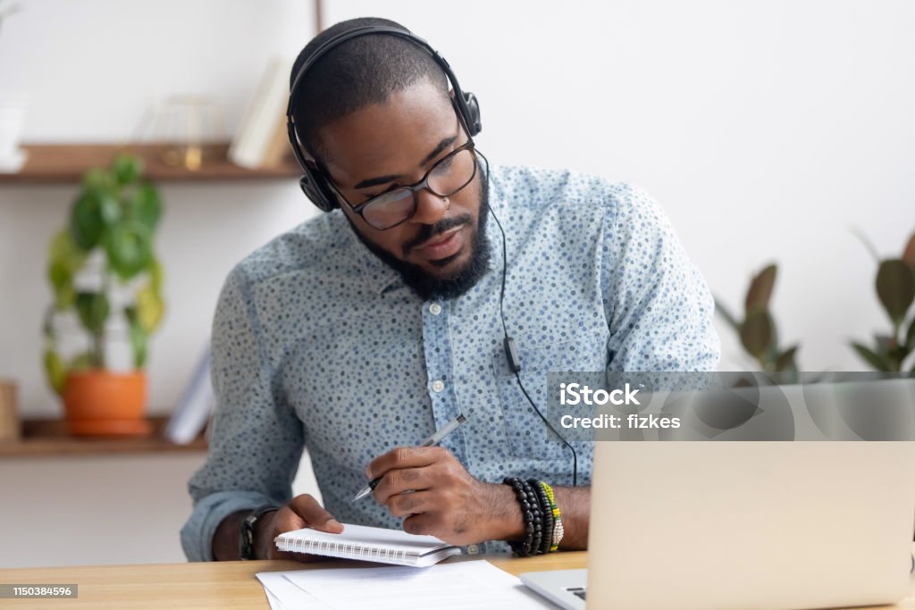 Focused african businessman in headphones writing notes watching webinar Focused african business man in headphones writing notes in notebook watching webinar video course, serious black male student looking at laptop listening lecture study online on computer e learning Learning Stock Photo
